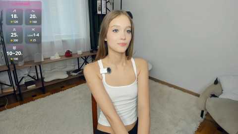 Video of a young Caucasian girl with straight light brown hair, wearing a white tank top and a microphone headset, sitting in a modern, minimalist living room.