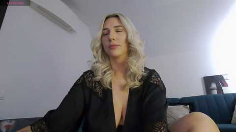 A video of a blonde, fair-skinned woman with wavy hair, wearing a black lace robe, sitting in a dimly lit room.