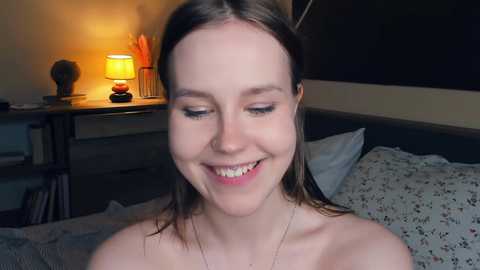 Video of a smiling young woman with straight, shoulder-length brown hair, wearing a strapless top, sitting on a bed with floral sheets, in a dimly lit room with a warm-lit lamp and plant on a bedside table.