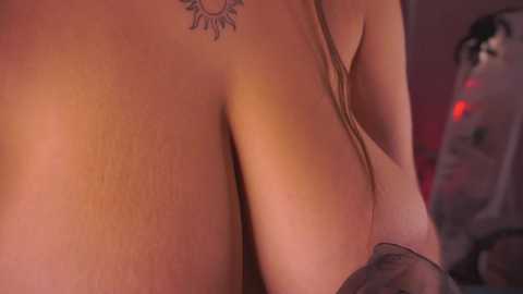 Video of a topless woman with large breasts and a tattoo of a sun near her neck, her skin tone is light, and she wears sheer black lingerie.
