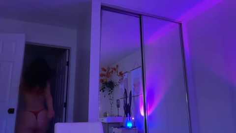 Video of a woman with dark hair, wearing a red bra, standing in a dimly lit bathroom with purple lighting. A large mirror reflects her and the room's interior.