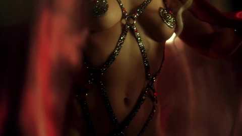 Video of a light-skinned woman with medium-sized breasts, wearing intricate, metallic bondage harnesses over her chest and hips. The background is dark and blurred, creating a dramatic, intimate atmosphere.