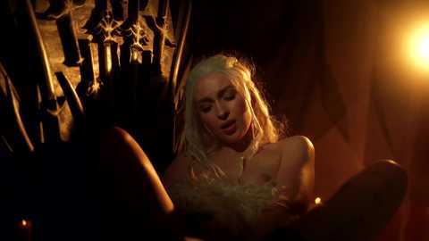 Video of a blonde woman in a dimly lit, medieval-style room, wearing a sheer, lace bodice, with a faint glow illuminating her face.