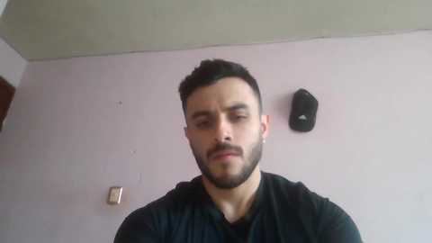 Video of a bearded man with short dark hair, wearing a black shirt, standing against a pale wall with a black speaker and a light switch.