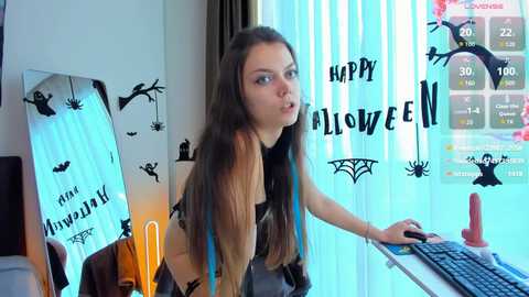 Media: Video of a young woman with long brown hair, dressed in black, playing a video game on a desk. The background features Halloween decorations and a \"Happy Halloween\" message.