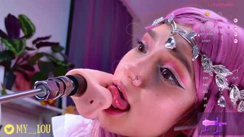 A close-up video of a young woman with light skin, pink hair, and silver jewelry, including a nose ring and headpiece, wearing a silver crown. She is seductively licking a metallic phallus. The background features a blurred potted plant and purple curtains.