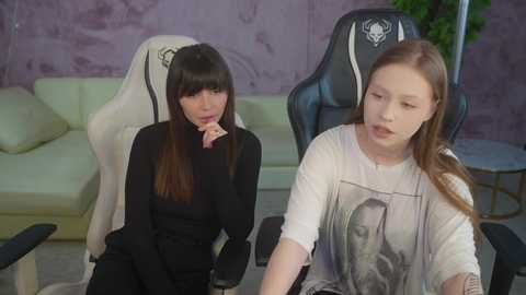 Video of two women in a gaming room: one with long brown hair in a black top, the other with long red hair in a white shirt with a graphic.