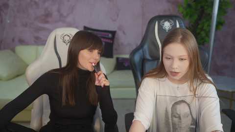 Media: Video of two young women, one with long straight brown hair, wearing a black turtleneck, sitting on a white gaming chair, the other with long blonde hair, wearing a white t-shirt with a monochrome portrait, sitting on a black gaming chair.