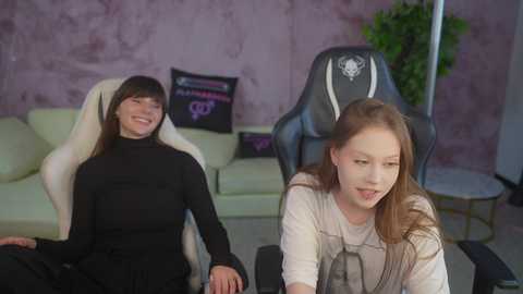 Video of two young women, one with straight brown hair, wearing a black turtleneck, sitting on a white gaming chair, and another with long red hair, wearing a beige sweater, seated on a black gaming chair, in a room with a purple wall and a potted plant.