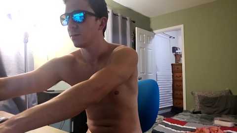 Video of a shirtless, slim, light-skinned man with glasses, blue reflective lenses, adjusting a camera in a cluttered, dimly lit bedroom with green walls, white door, and a blue chair.