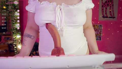Video of a light-skinned woman wearing a white off-the-shoulder dress with a large, erect, realistic pink dildo inserted into her vagina. The background features pink walls, fairy lights, and a poster.