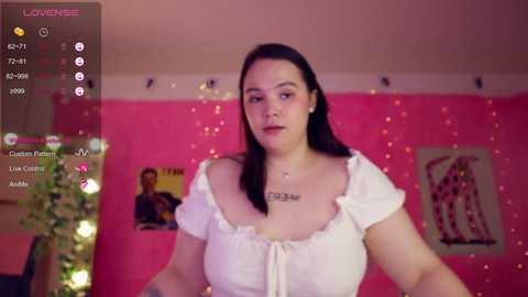 Video of a fair-skinned woman with long dark hair, wearing a white off-shoulder blouse, in a pink room with string lights and artwork.