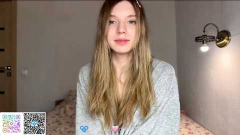 Video of a young Caucasian woman with long, straight, blonde hair, wearing a light grey cardigan, standing in a simple, softly lit bedroom with floral-patterned bedding.