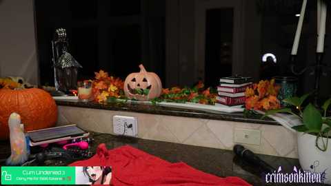 Media: Video of a Halloween-themed kitchen countertop with a carved pumpkin, orange leaves, books, candles, and a red cloth, set against a dark background.
