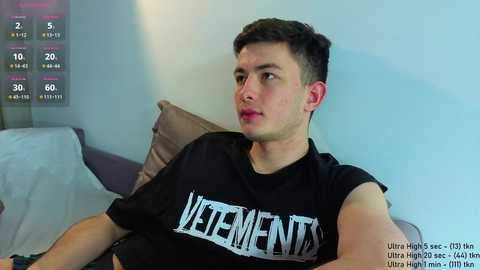 A video of a young, fair-skinned man with short brown hair, wearing a black \"VETEMENTS\" t-shirt, sitting on a bed with a white and purple duvet, against a light blue wall.