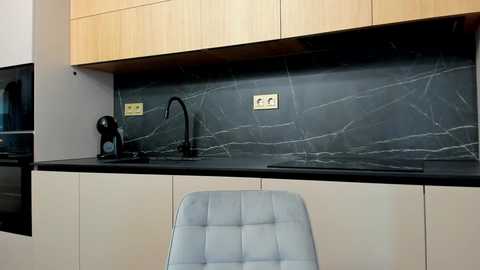 Video of a modern kitchen with light wooden cabinets, dark gray marble backsplash, stainless steel appliances, and a light blue, tufted chair.