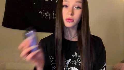 Video of a young woman with long brown hair, wearing a black t-shirt, holding a black umbrella with white graffiti. She has a serious expression, and the background is a plain beige wall.