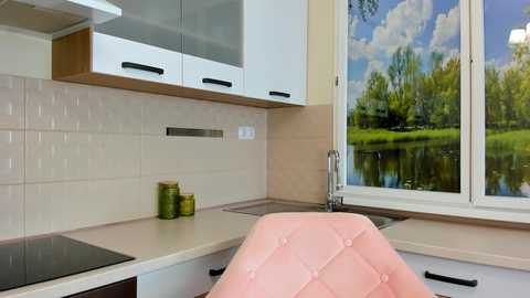 Video of a modern kitchen with white cabinets, beige tiles, a pink chair, and a large window showing a serene outdoor scene.