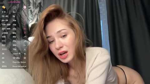 A video of a fair-skinned, blonde woman with long hair, wearing a beige top, lying on a bed, mouth open, surrounded by dark curtains, and a virtual reality headset.