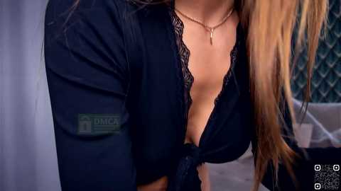 Video of a woman with long, blonde hair, wearing a black lace-trimmed blouse tied at the waist, revealing cleavage, and a gold necklace. Background shows a chain-link fence and blurred furniture.