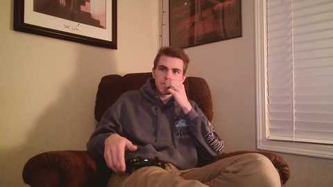 Video of a young man in a gray hoodie, sitting on a brown recliner, with a remote in hand, looking contemplative, in a beige room with framed art on the wall and closed blinds.