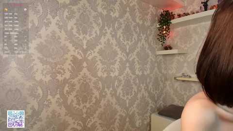 Media: Video of a woman with a short, dark bob haircut, viewed from the back, standing in a room with a grey, floral-patterned wallpaper.