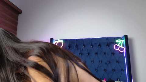 Video of a woman with long, dark hair and bare back, standing in front of a tufted, dark blue headboard with glowing neon cherries and a pink heart, set against a plain white wall.