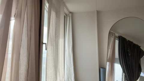 Video of a minimalist, well-lit room with beige walls and sheer, light-colored curtains partially drawn over a window. A large, round mirror reflects part of the room, adding depth.
