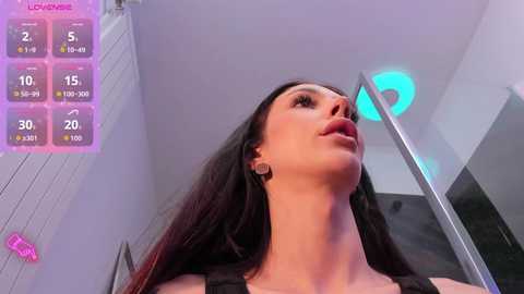 Video of a young woman with long, dark hair, wearing a black tank top, looking upward in a modern, white-walled room with a blue neon light.