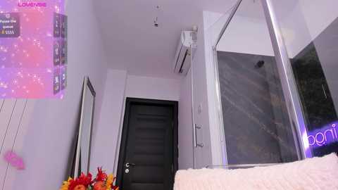 A video of a modern, minimalist bedroom with a black door, white walls, an air conditioning unit, and a glass shower door. A pink light display adorns the wall, and a plush beige sofa is visible in the foreground.