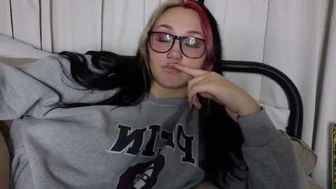 Video of a young woman with long black and red hair, wearing glasses and a grey sweatshirt, lying on a bed, fingers to her lips, in a dimly lit room with white curtains.