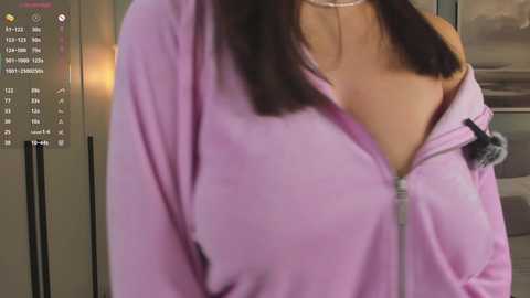 A video of a woman with medium skin tone, wearing a pink zip-up hoodie with the zipper partially open, revealing a black bra. The background shows a modern kitchen with stainless steel appliances.