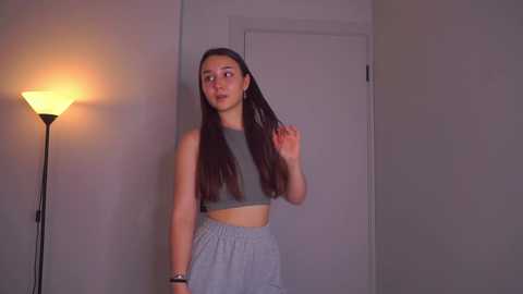 Video of a young woman with long brown hair, wearing a gray crop top and high-waisted pants, standing indoors, adjusting her hair, illuminated by a nearby lamp.