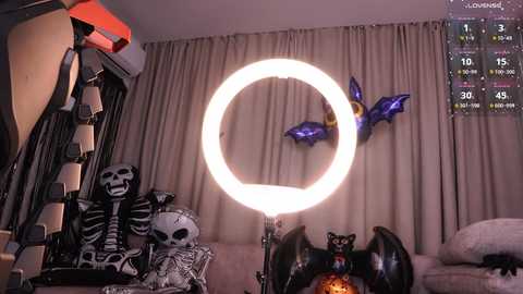 Video of a Halloween-themed living room with a glowing ring light, black bat decorations, and a skeleton plush on a beige sofa.