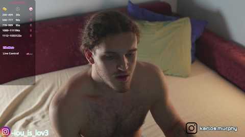 A video shows a shirtless, fair-skinned man with curly brown hair, sitting on a bed with red and blue pillows. The image has social media icons and a live stream overlay.