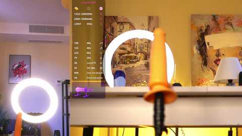 Video of a cozy, well-lit room with a white mirror, two large round lamps, a clock, and a bright orange dildo on a white table.