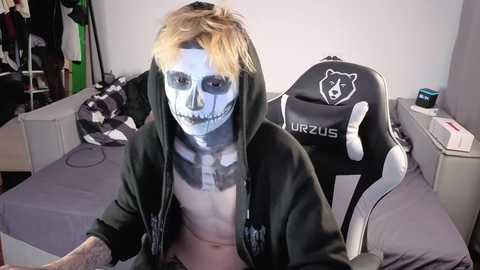 Media: Video of a person dressed as a skeleton with a black hoodie, sitting in a gaming chair with a \"Urza's\" logo, in a cluttered, dimly lit room with clothes and gaming equipment.