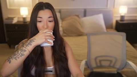 Video of an Asian woman with long black hair, tattoos on her arm, drinking from a white cup in a beige room with a bed, nightstands, and a chair.