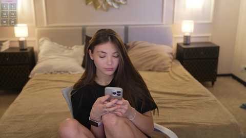 Video of a young woman with long dark hair, sitting on a bed, holding a smartphone, in a beige room with two lamps and a floral artwork.