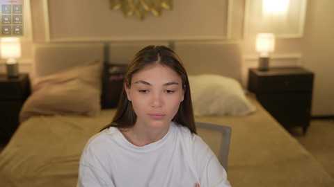 Video of a young woman with straight, dark hair, wearing a white t-shirt, sitting in a cozy bedroom with beige walls, a bed, and two nightstands with lamps.