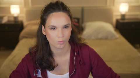 Media: Video of a young woman with medium brown skin, dark hair, and a maroon jacket, standing in a dimly lit bedroom with a bed, two nightstands, and lamps.