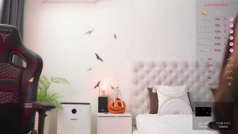 Video of a modern, minimalist bedroom with a tufted gray headboard, a small white table with a pumpkin candle, a black gaming chair, and a calendar with a pink background.