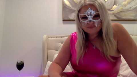 Video of a curvy, blonde woman with a large chest in a pink one-shoulder dress, wearing a silver masquerade mask with fringe. She's on a bed with white bedding and a beige headboard, in a softly lit room with a framed painting on the wall.