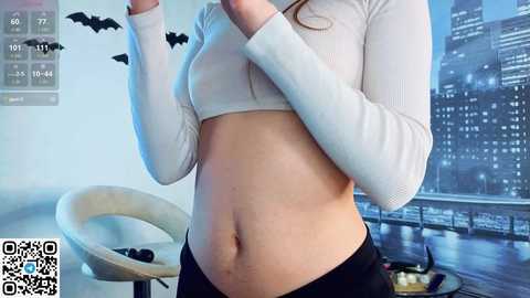 Media: Video of a fair-skinned woman with long hair, wearing a white, long-sleeved crop top and black pants, in a modern living room with a cityscape mural and bat decorations on the wall.