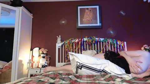 Video of a woman lying naked on a bed, wearing a black bra, in a cozy, maroon-walled bedroom with a white crib, colorful hanging toys, and a framed picture on the wall.
