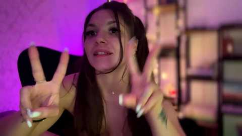 Video of a young woman with long brown hair, smiling and making peace signs, in a dimly lit room with purple and pink lighting, black furniture, and shelves in the background.