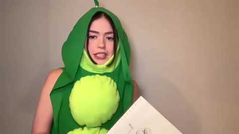 Media: A young woman with light skin and dark hair wears a green, plushie dragon costume with a hood and large, round, green belly. She holds a piece of paper, smiling. Plain beige background.