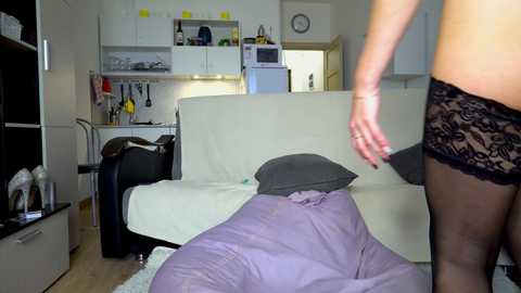 Video of a woman in black lace panties and thigh-high stockings, standing over a man sleeping on a white sofa in a modern kitchen.