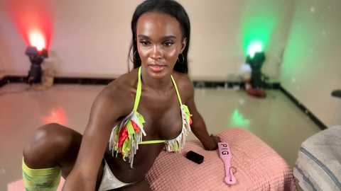 Video of a dark-skinned woman with long black hair, wearing a neon-green bikini with colorful fringe, sitting on a pink quilted cushion in a studio with green and red stage lights, black floor, and a pink bracelet.