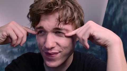 Video of a young man with wavy brown hair, smiling, pointing at his eyes, wearing a black shirt, against a blurred green forest background.